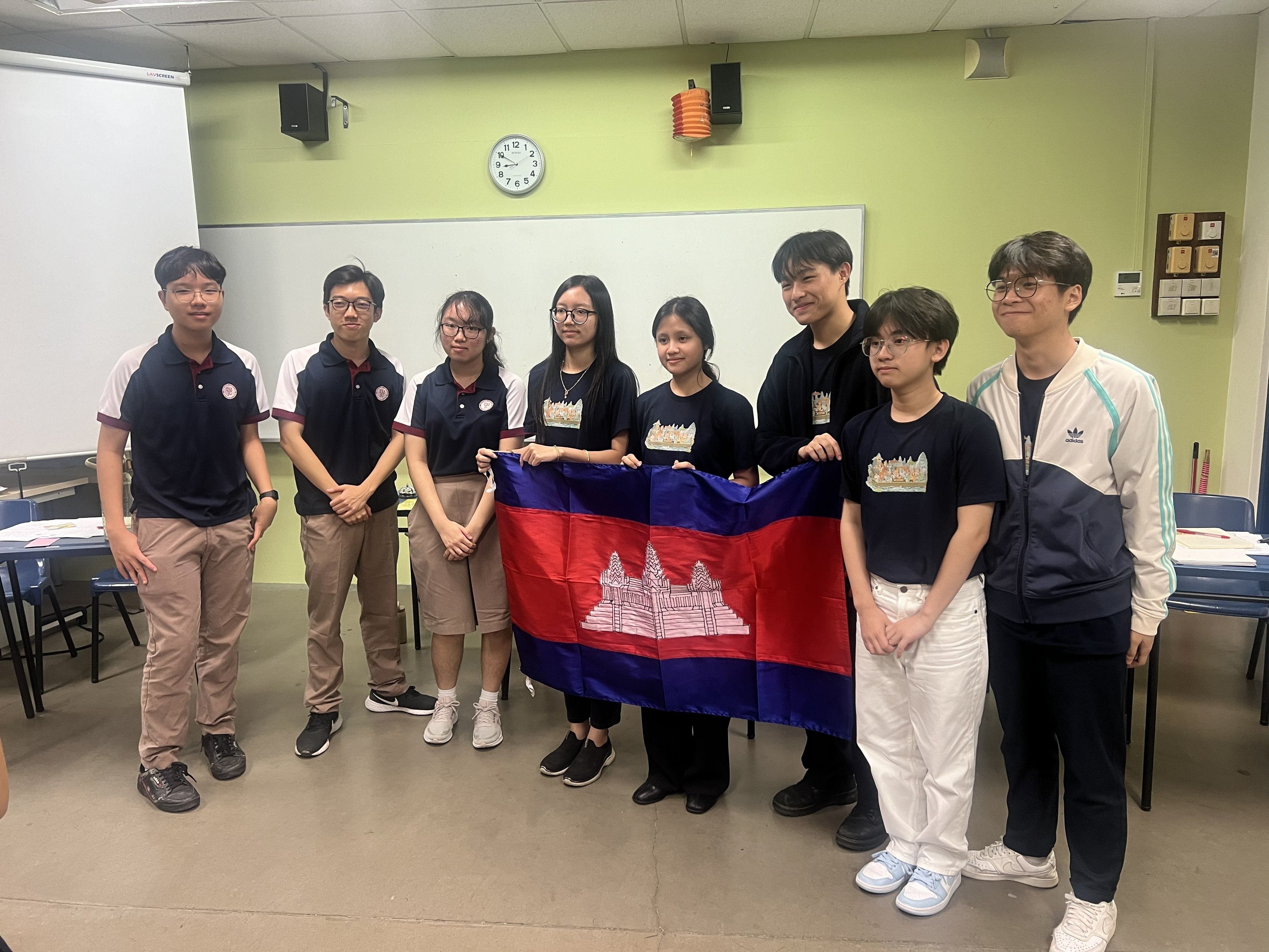 Eight students stand in a row inside a classroom, posing for a group photo. Two students in the centre hold up the Cambodian flag, which is blue and red with an image of Angkor Wat in the middle. Some students are in TMJC school uniforms while others wear matching black T-shirts with a printed design. A whiteboard, clock, and green walls are visible in the background.
